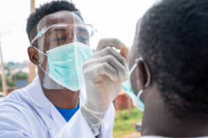 Male medical worker in full protective gear administering a COVID-19 nasal test to a male patient outdoors.
