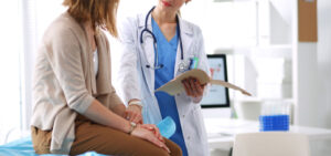Female doctor holding chart and discussing something with female patient sitting on examination bed.