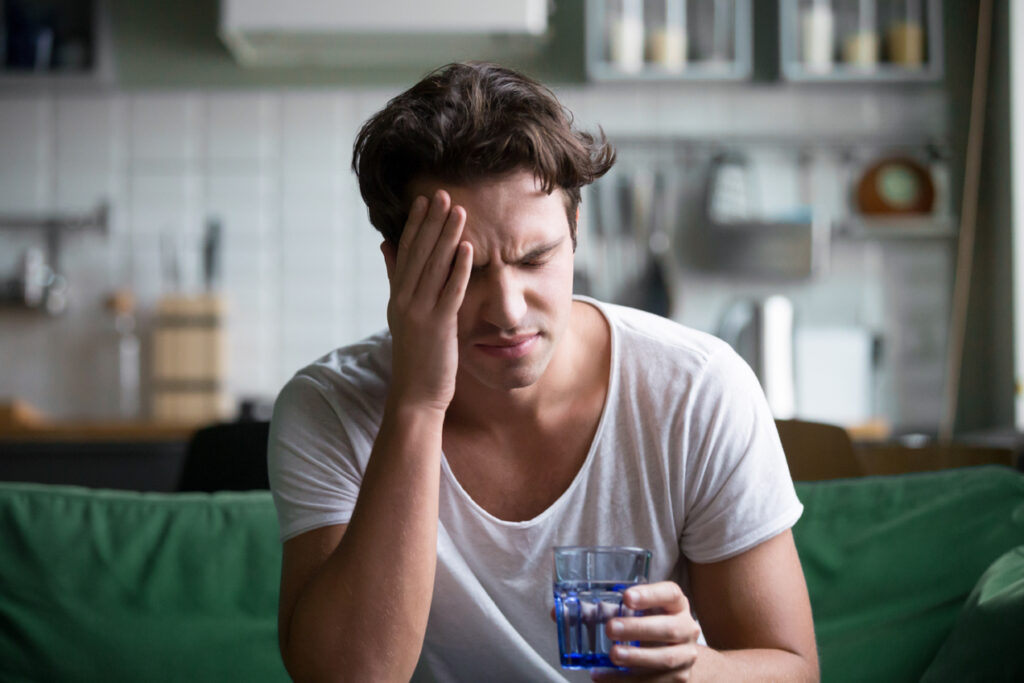 White male sitting on the sofa holding a glass of water with his palm against the side of his forehead with a look of pain on his face.