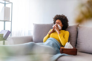 Sick woman sitting on a sofa covered in a blanket with a box of tissues by her side blowing her nose.