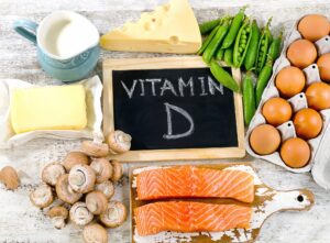 A chalkboard on a rustic table with the word “Vitamin D” on it surrounded by eggs, milk, cheese, butter, peas, salmon, and mushrooms.