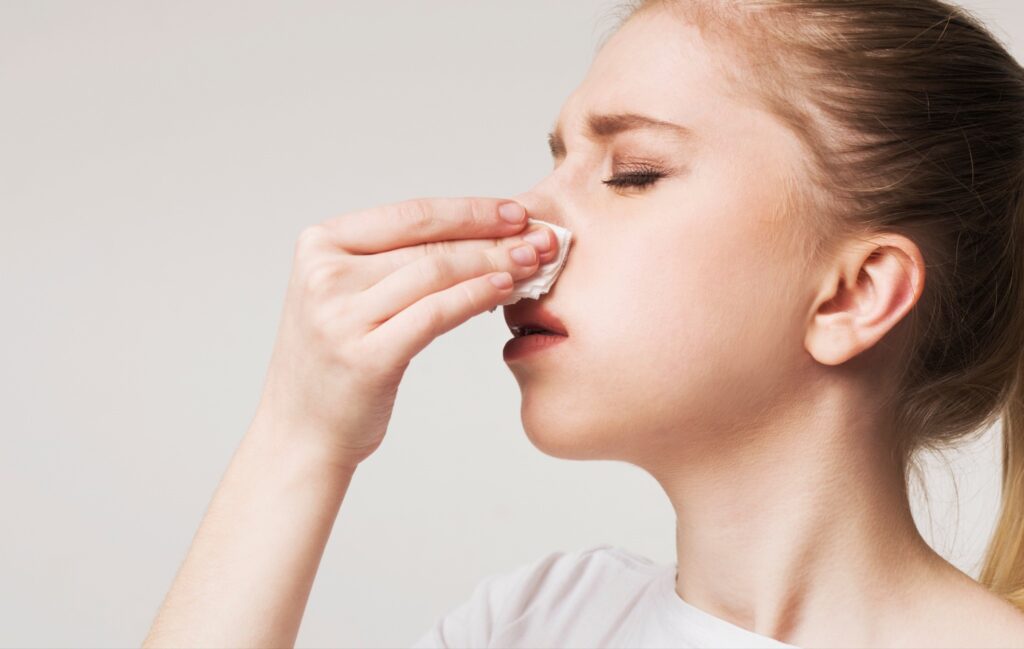 A closeup of a woman tilting her head back slightly while holding a tissue to her nose with her eyes closed.