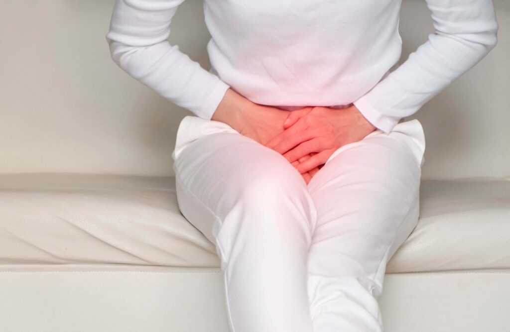 A close-up image of a woman sitting on a white couch with both hands over her lower abdomen.
