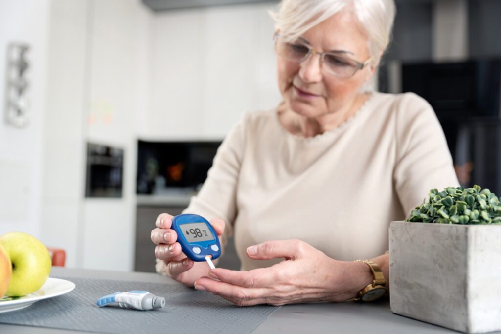 An older woman is sitting at her kitchen table checking her blood sugar with a blood test monitor.