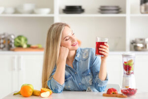 A blonde woman sitting at a countertop in her kitchen next to a blender and fruit, smiling at a glass of fresh cranberry juice in her hand.