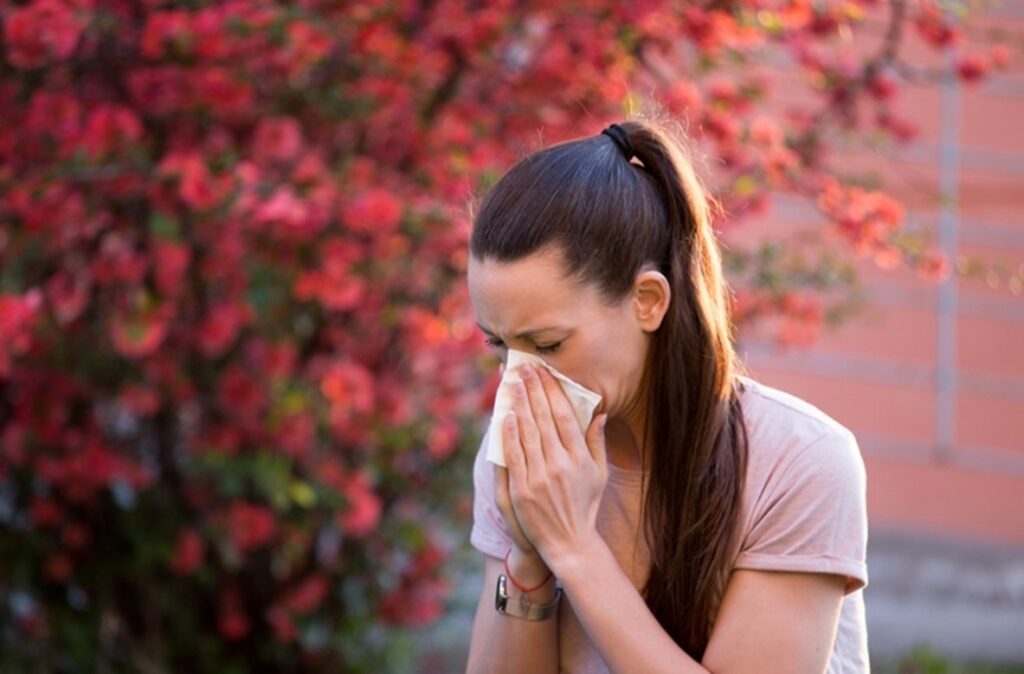 A woman is standing outside near a blossomed tree, blowing her nose into a tissue.