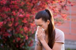 A woman is standing outside near a blossomed tree, blowing her nose into a tissue.