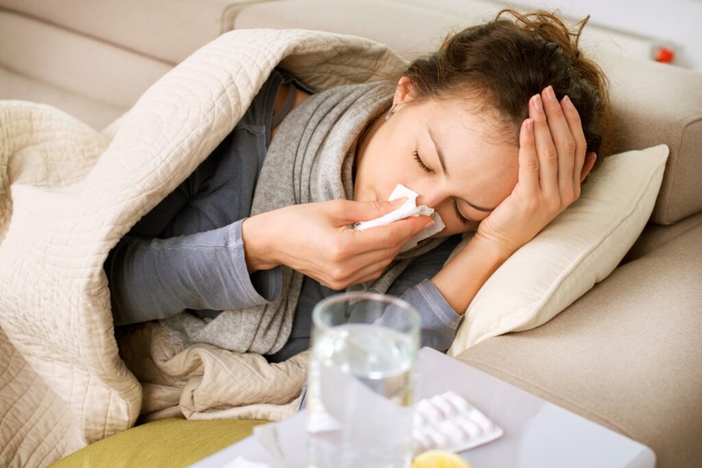 A sick woman lying on the sofa covered in a blanket and blowing her nose next to a table with a cup of water and medicine on it.