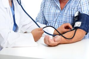 A close-up image of a doctor in a white coat with a stethoscope checking the blood pressure on the arm of a male patient.
