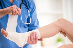 A closeup image of a doctor in blue scrubs with a stethoscope around his neck wrapping the hurt ankle of a patient.