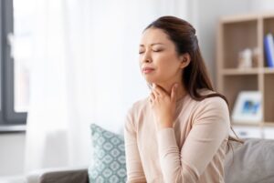 A woman sits on her sofa with her eyes closed and a hand resting against her sore throat.