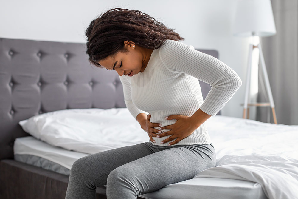A closeup of a woman sitting on the edge of her bed, gripping her abdomen in pain with both hands.