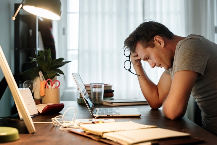 A middle-aged man stares at his computer screen on his messy desk while holding his glasses in his hands with a stressed-out look on his face.