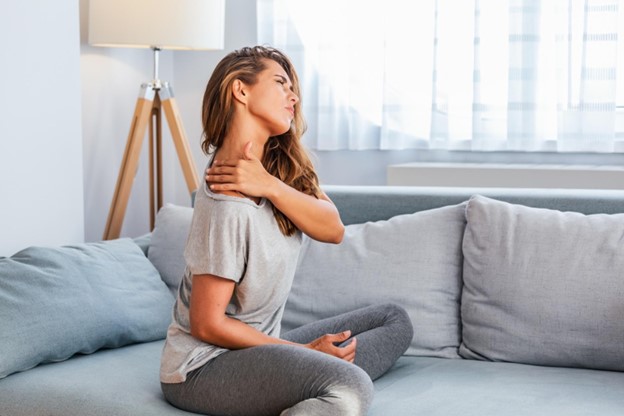 A woman sits on her sofa, tilting her head and rubbing her neck with one hand while wincing in pain.