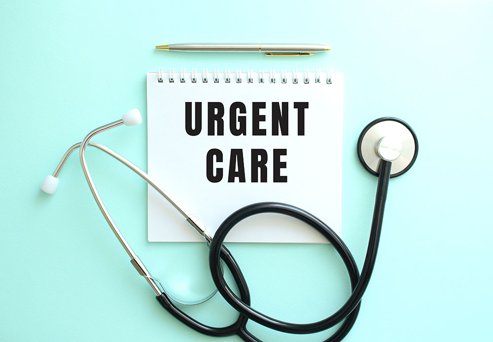 A stethoscope lays across a white notepad with the words “urgent care” written on it. Above the notepad is a pen.