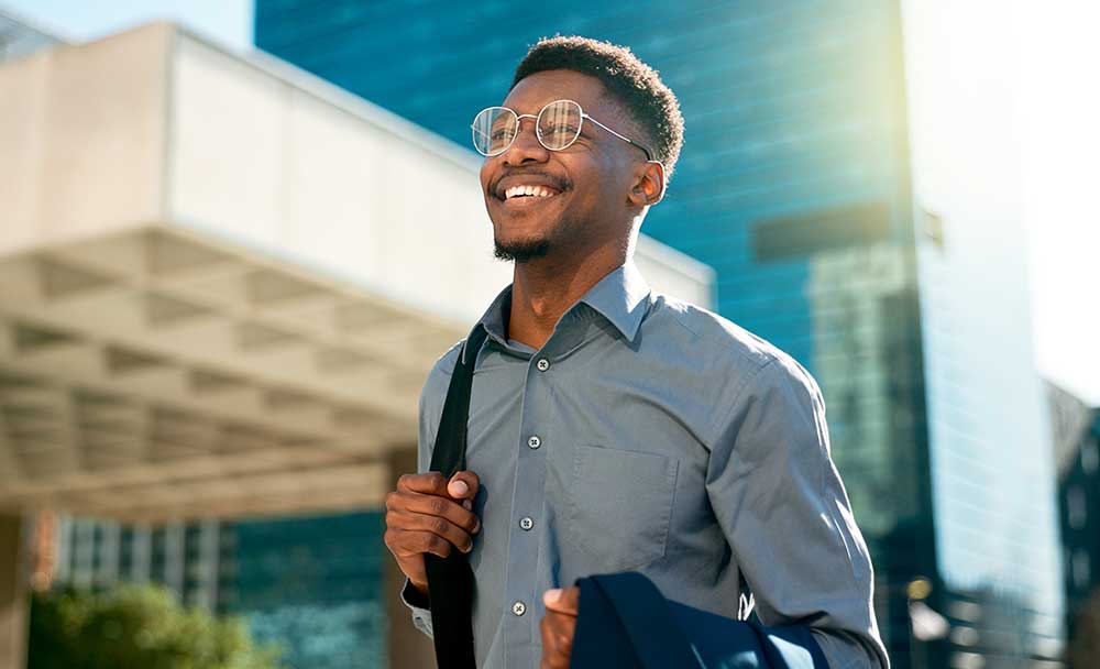 A man wearing glasses and carrying a bag over his shoulder is smiling and walking down a city street.