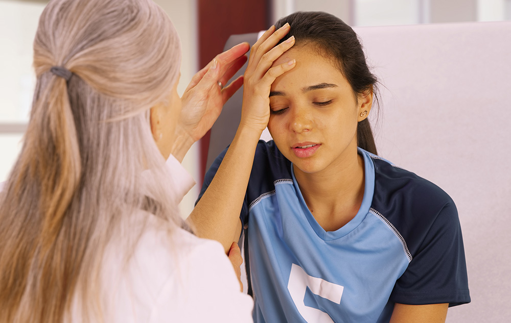 A girl wearing a soccer jersey holds her right hand to her head. A woman in a white lab coat sits in front of her and reaches out to do an assessment.