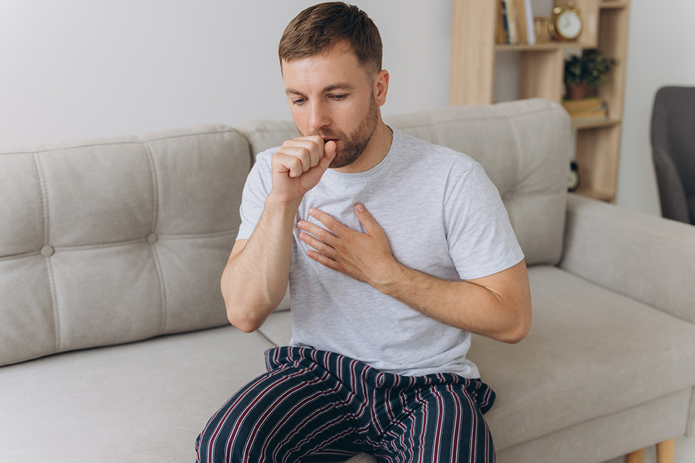 A man sitting on a couch holds his left hand to his chest and his right hand to his mouth. He appears to be coughing.