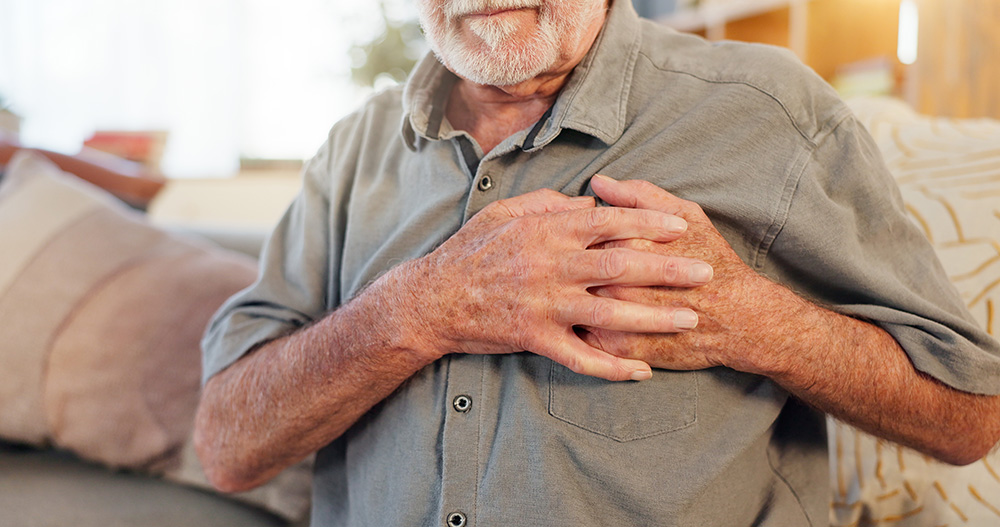 A man sitting on a couch clutches the left side of his chest with both hands.