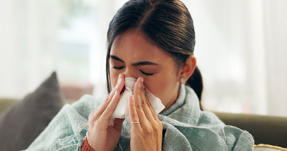 A closeup image of a woman wrapped in a blanket blowing her nose into a tissue.