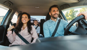 A man and a woman sit in the front seats of a car with a girl in the backseat. It is sunny outside and all three are smiling.