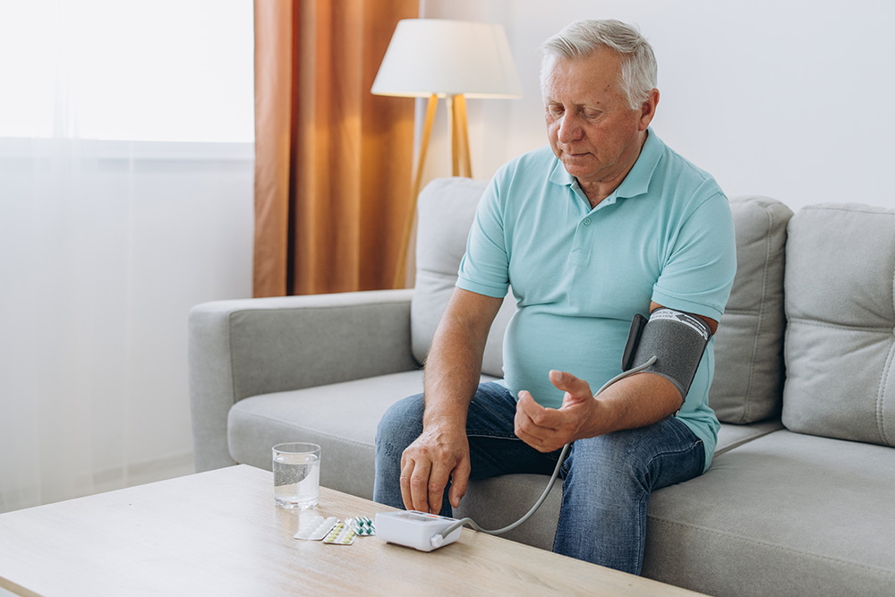 An older man sits on a couch with a blood pressure cuff around his arm. A glass of water and medication are on a table in front of him.