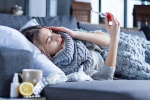 Sick woman lying in bed wrapped in blanket and scarf, holding thermometer to check temperature while treating cold or flu at home.