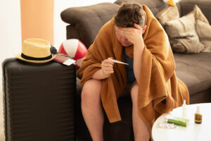 Sick man covered by a blanket looking at a thermometer. He is surrounded by a suitcase,